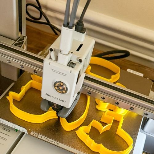 A close-up, high-angle shot shows a Bambu Lab 3D printer in action, extruding bright yellow filament onto a metallic print bed. Several partially formed and complete cookie cutters, also in bright yellow, are visible on the bed, including shapes like a banana and a flower. The printer's print head, a white rectangular device with a nozzle at the bottom, is centered in the frame, actively creating one of the shapes. Wires extend from the top of the print head, and the printer's gantry system is visible, indicating the print head's movement across the bed.