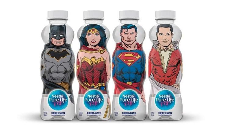 Four Nestle Pure Life bottled waters, each shaped and decorated like a different DC Comics superhero, stand in a row against a plain white background. From left to right, the bottles feature Batman in a grey and black suit, Wonder Woman in a red and gold outfit, Superman in his classic blue and red costume, and Shazam in a red and white suit. Each bottle has a white cap and a small label at the bottom with the Nestle Pure Life logo and the words 'Purified Water'.