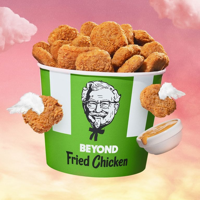 A vibrant, surreal image features a large green and white KFC bucket overflowing with golden-brown Beyond Fried Chicken nuggets. The bucket has a smiling Colonel Sanders logo and the text "BEYOND Fried Chicken" in black. The background is a dreamy pink and orange sky with fluffy white clouds. Two nuggets with small white wings float on either side of the bucket, and a small white bowl of dipping sauce with a nugget floating above it is positioned in the bottom right corner.
