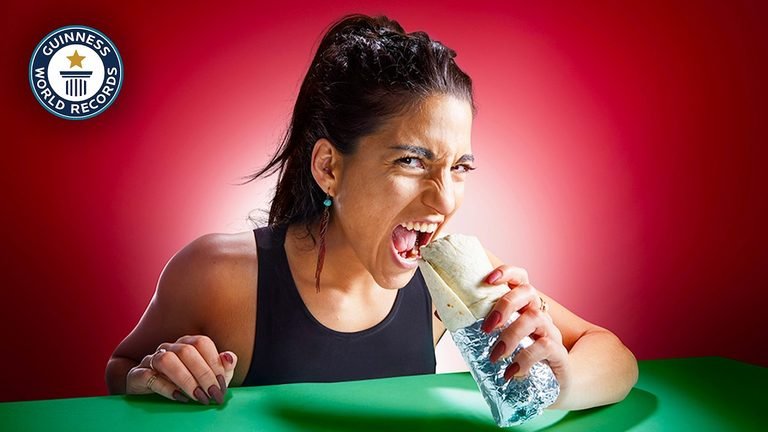 A woman with dark hair pulled back and wearing a black tank top takes an aggressive bite from a large, foil-wrapped burrito. Her face is contorted in an intense, almost fierce expression with wide eyes and an open mouth, as she sits at a green table against a vibrant red background, with the Guinness World Records logo in the top left.