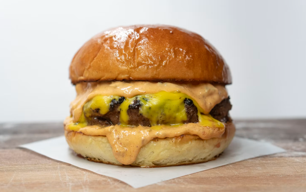 A close-up, eye-level shot of a delicious-looking cheeseburger with a glossy, toasted bun. The burger features two patties covered in melted yellow cheese and a generous amount of creamy, orange-pink sauce that drips down the sides onto the bottom bun. It rests on a small piece of white parchment paper on a light wooden surface, with a plain white background.