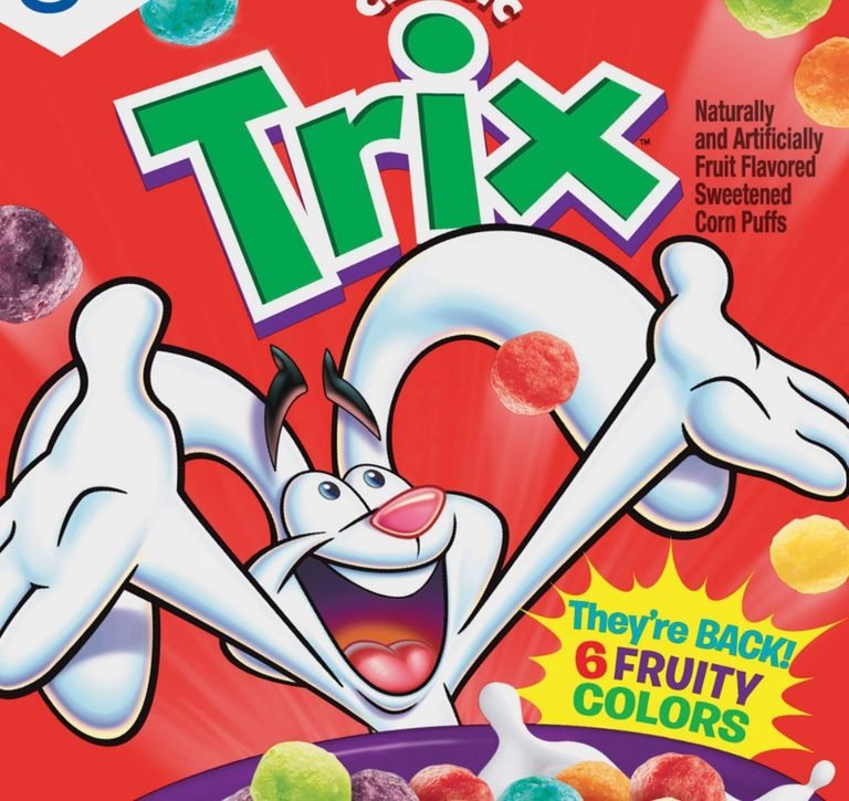 A vibrant red Trix cereal box features the Trix Rabbit, a white cartoon rabbit with black ears and a pink nose, excitedly reaching out with both hands as colorful, spherical Trix cereal pieces float around him. The large green and purple 'Trix' logo is prominent at the top, with text to the right stating 'Naturally and Artificially Fruit Flavored Sweetened Corn Puffs.' A yellow starburst near the bottom right announces 'They're BACK! 6 FRUITY COLORS,' with several cereal pieces visible in a purple bowl with milk.